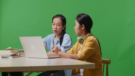 asian woman students arguing about something on the laptop while sitting on a table studying in the green screen background classroom
