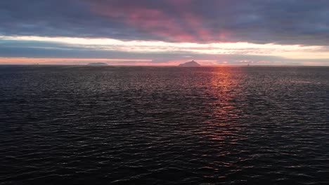 Slowly-flying-sideways-over-a-calm-ocean,-with-islands-and-a-cloudy-sunrise-on-the-horizon