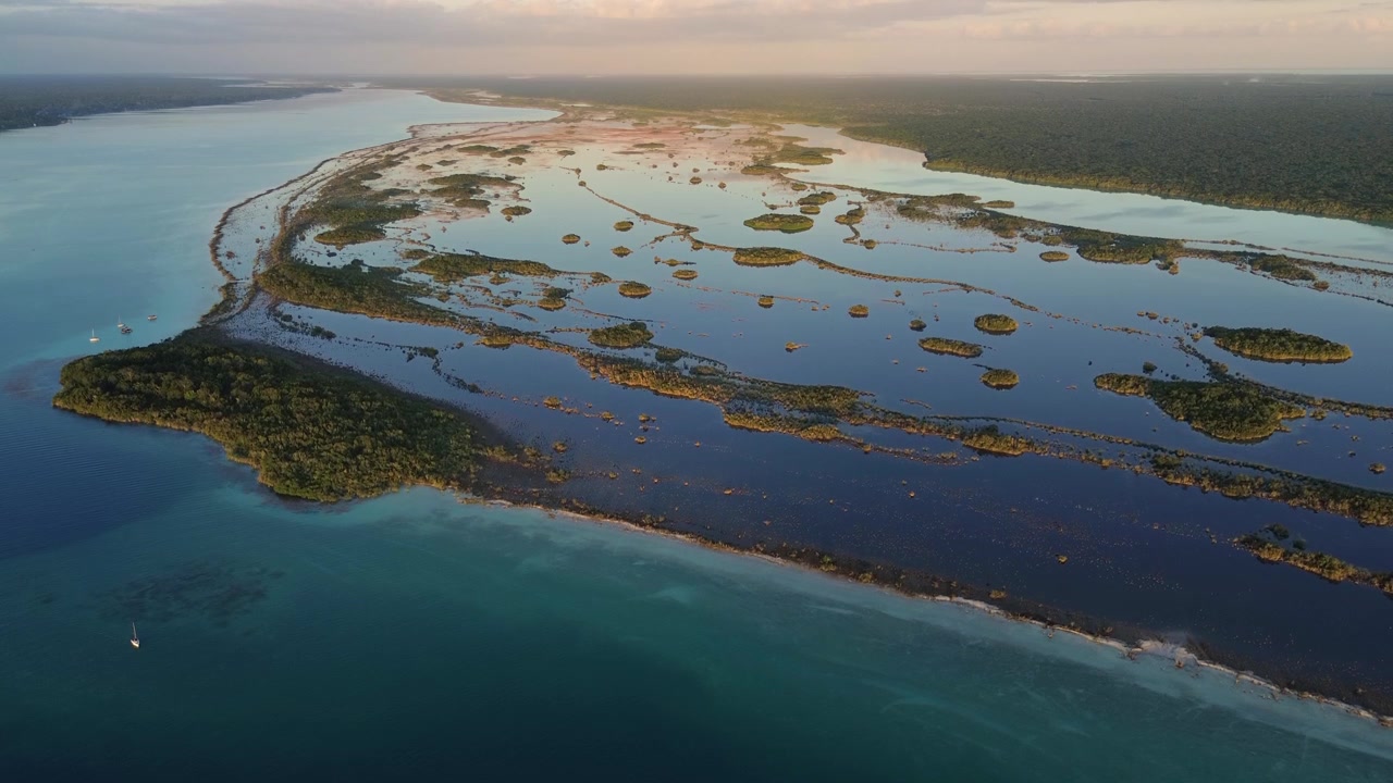 Tomada de drone ascendendo sobre a vasta Lagoa de Bacalar, revelando padrões abstratos de manguezais e um veleiro solitário cruzando águas azuis calmas no México.