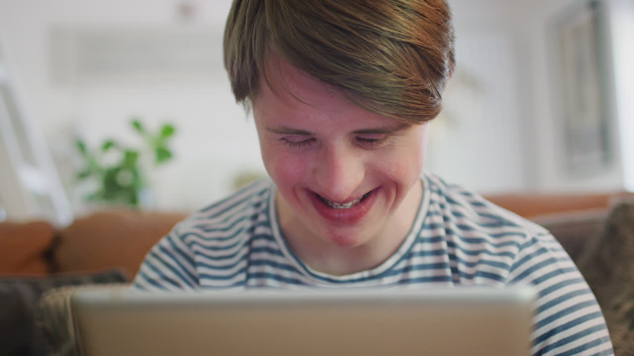 젊은 다운 증후군 남자가 소파에 앉아 집에서 디지털 태블릿을 보고 있습니다.