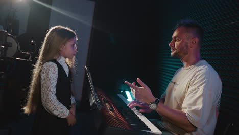 blonde child and trainer work in music studio. man plays synthesizer while little girl does vocal warm up in sound insulated room. singing lesson