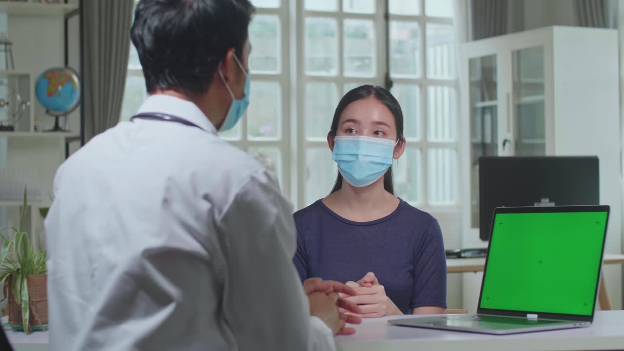 Asian Man Doctor Showing Green Screen Display On Laptop Computer During Consultation In A Health Clinic. Both Wear Face Masks. Physician In Lab Coat Sitting Behind A Computer Desk In Hospital Office