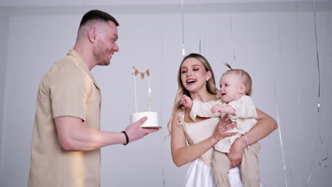 loving caucasian parents congratulating their beloved baby with first birthday. father is holding a cake and mother blows the candle.