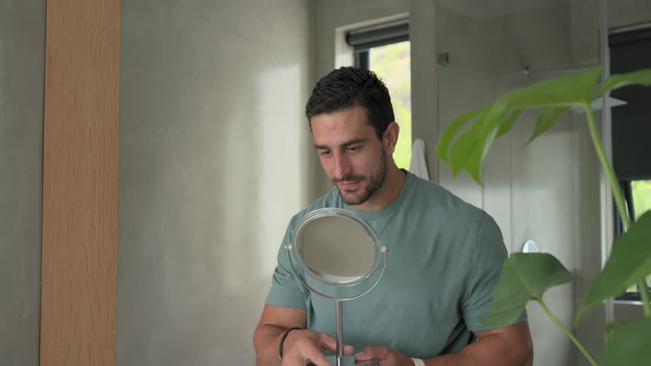 Uomo adulto che afferra lo specchio davanti allo specchio, sollevandolo e inclinandolo, controllando guance e collo per la barba incolta