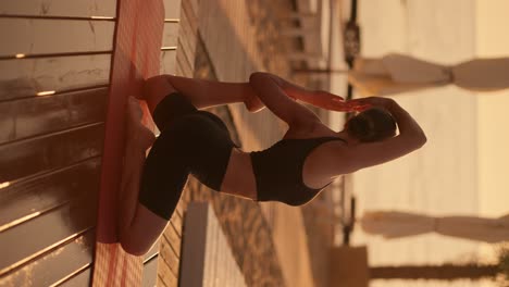 vertical video: a brunette girl with tied hair in a black sports summer uniform takes a special stance during a yoga class on a special red mat on a sunny beach during a golden sunset in summer