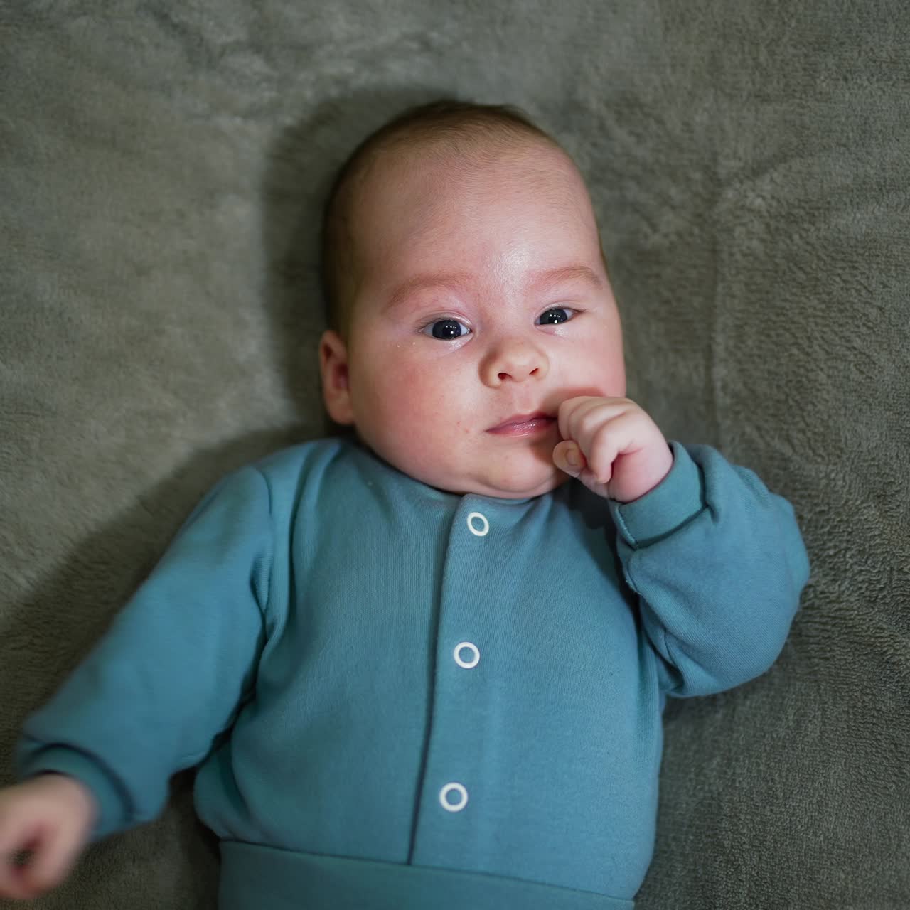 Baby boy wearing blue warm suit lying on the soft bed. Child moving his hands actively. Grey blanket background