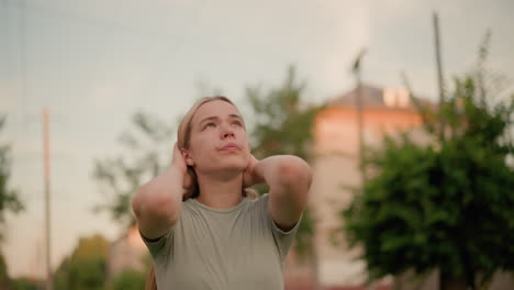 young woman outdoors with hands on her head, gazing upward with a contemplative, moody expression, surrounded by blurred greenery and urban structures