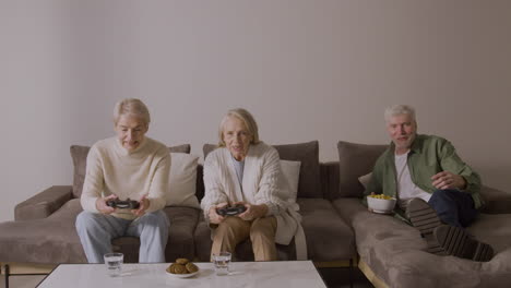dos ancianas felices jugando videojuegos sentadas en un sofá en casa mientras un anciano entusiasta tumbado en el sofá mirándolas y comiendo algo 2