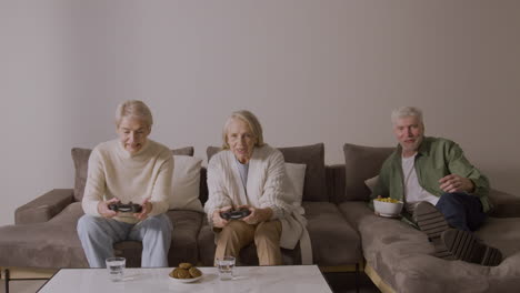 two happy senior women playing video game sitting on sofa at home while enthusiastic elderly man lying on sofa watching them and eating something 2