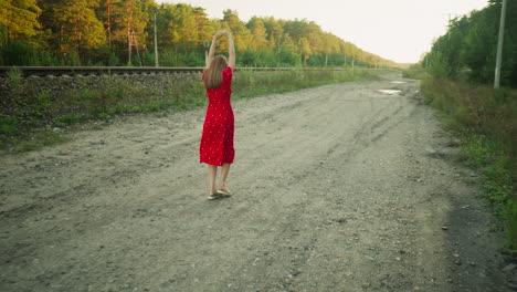 Woman in red dress dancing and walking joyfully along countryside dirt road beside railway track, surrounded by natural greenery and sunset light in relaxed outdoor rural environment