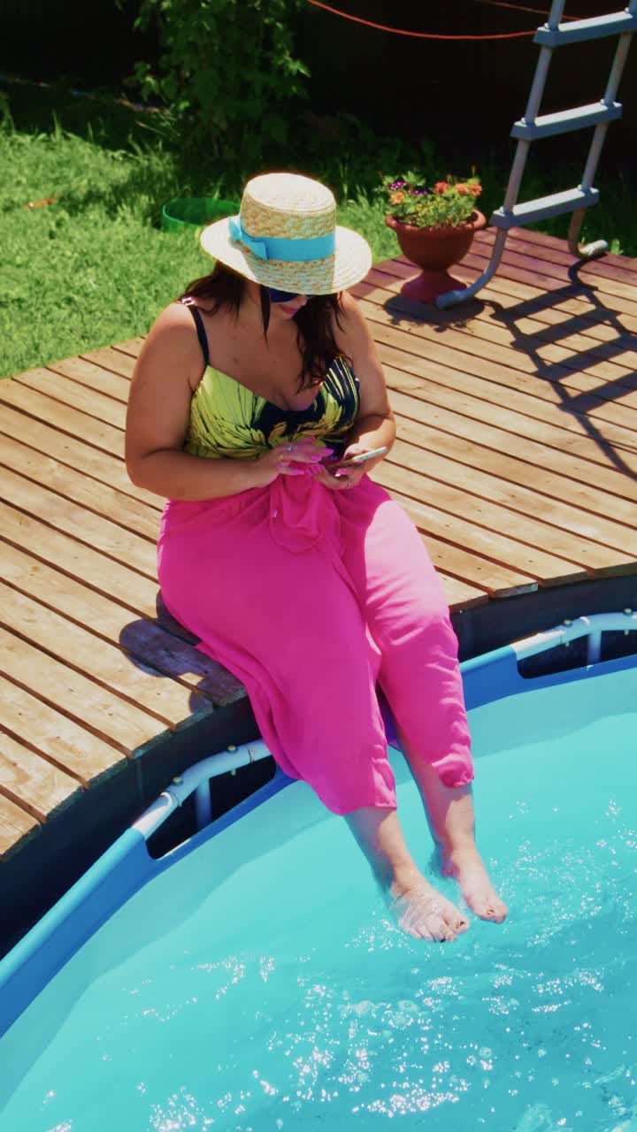 Relaxing by the Poolside: A Woman Enjoying a Warm Day in a Colorful Hat and Bright Cover-Up While Using Her Phone by the Tranquil Water Surface