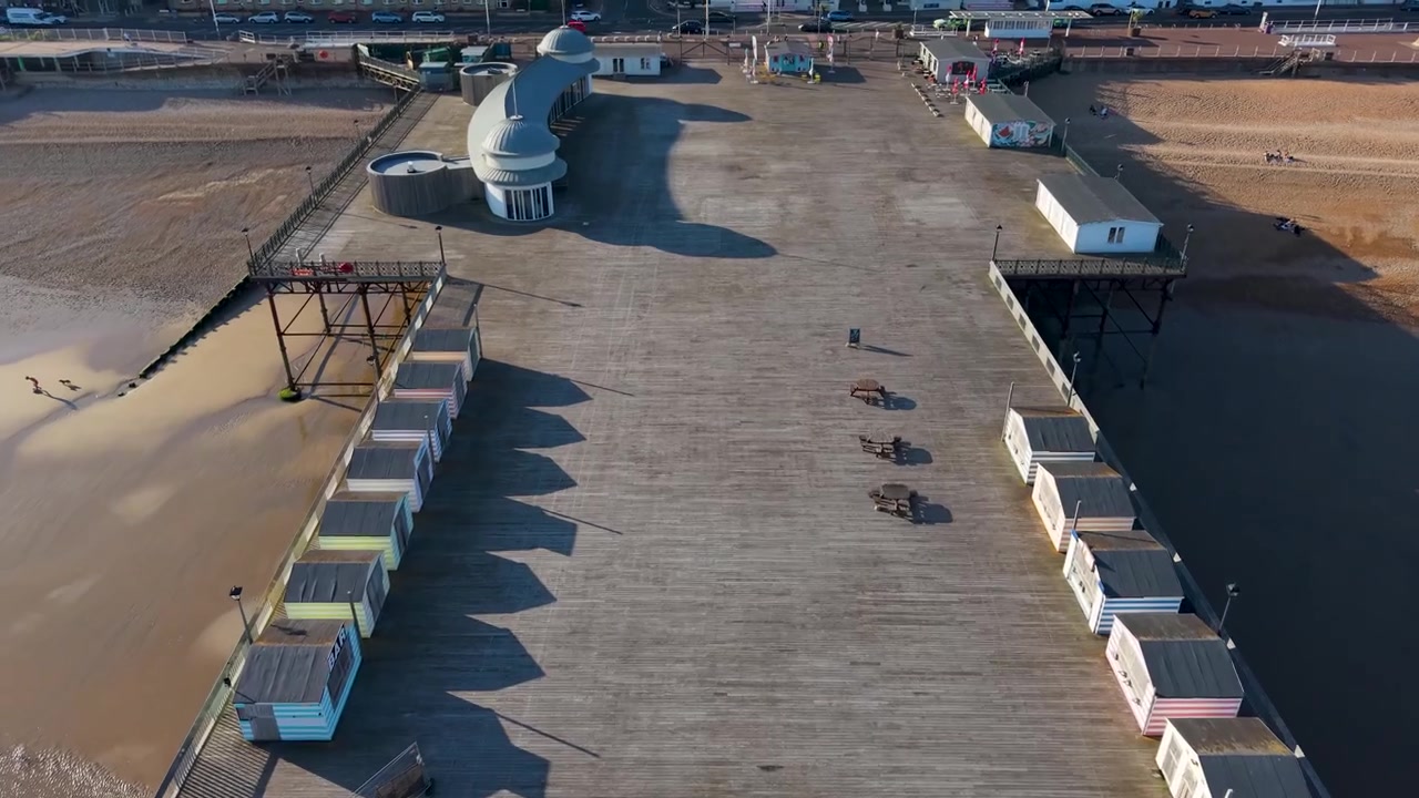 Un sobrevuelo rápido y bajo de dron recorre la cubierta del muelle de Hastings, pasando por la zona de asientos elevados y vestuarios, y el Pabellón Occidental curvo, antes de llegar a la carretera costera en la entrada.