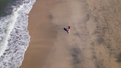 a drone follows a woman as she walks along a beach as seen from an aerial overhead drone