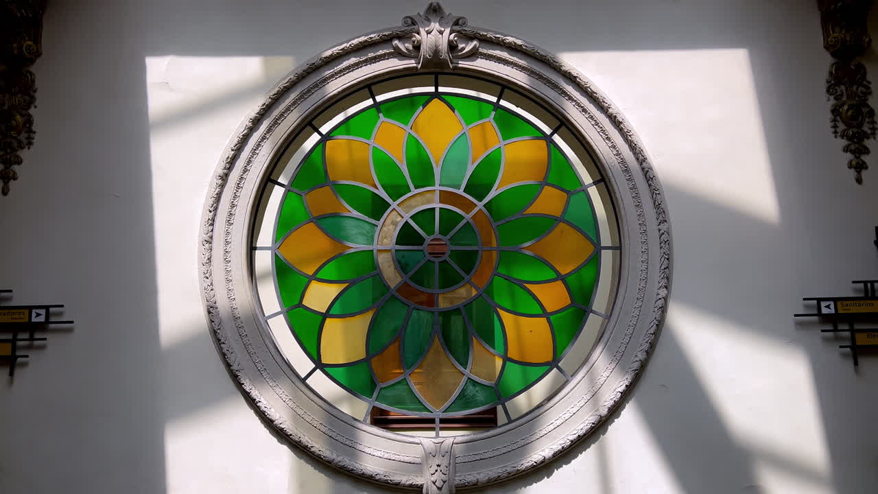 Primer plano de una vidriera circular con vibrantes diseños florales verdes y amarillos que exhiben una artesanía elegante y el juego de luz natural en un interior brasileño histórico