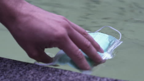 a simple green protective covid-19 face mask floating on restless water of a city fountain, thrown out, soaked and wet, being taken out of the water carefully by somebody, static close up 4k shot