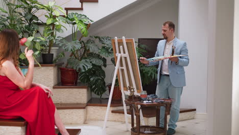 artist draws wife portrait with rose at home. beautiful woman in long red dress sits on podium while master works in residence. art studio work