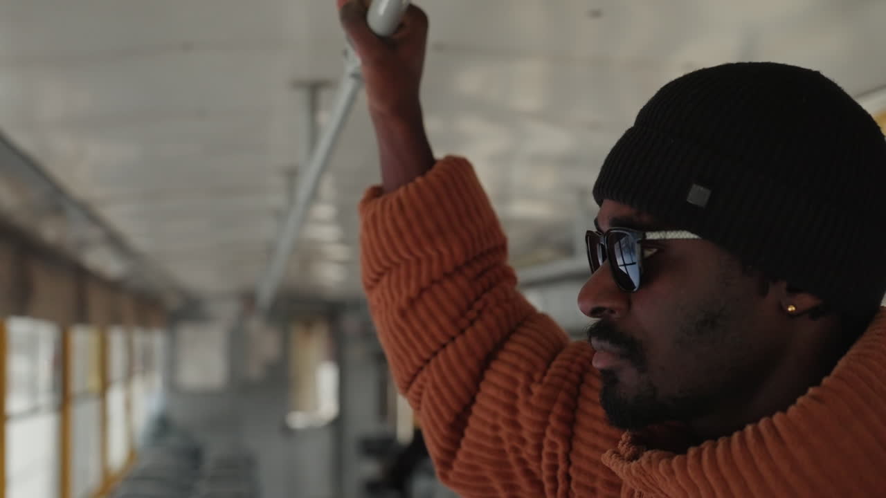 Black man gripping pole on bus at dusk, knit beanie and sunglasses, warm sweater against cold light, focused gaze toward window, industrial interior rail, blurred city lights outside, candid travel
