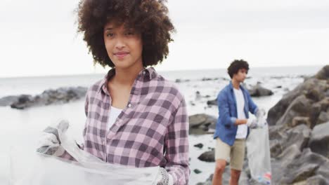 Porträt-Einer-Afroamerikanischen-Frau,-Die-Lächelt,-Während-Sie-Müll-Auf-Den-Felsen-Am-Meer-Sammelt