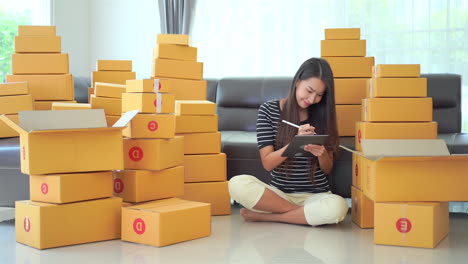 Negocio-Desde-Casa,-Joven-Asiática-Haciendo-Una-Lista-De-Entrega-En-Una-Tableta-Para-Enviar-Cajas-De-Paquetes-Desde-Su-Casa,-Trabajando-En-Casa