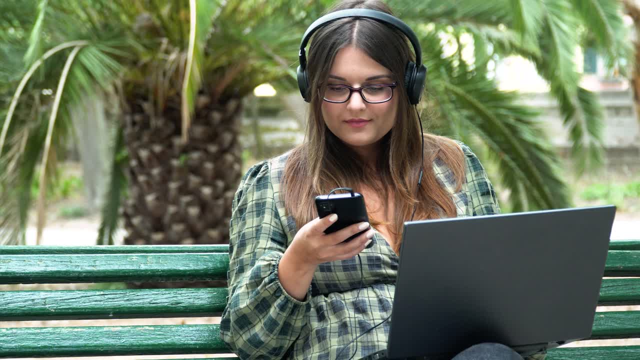 una hermosa chica con auriculares y una laptop en el parque está hablando por teléfono y riéndose