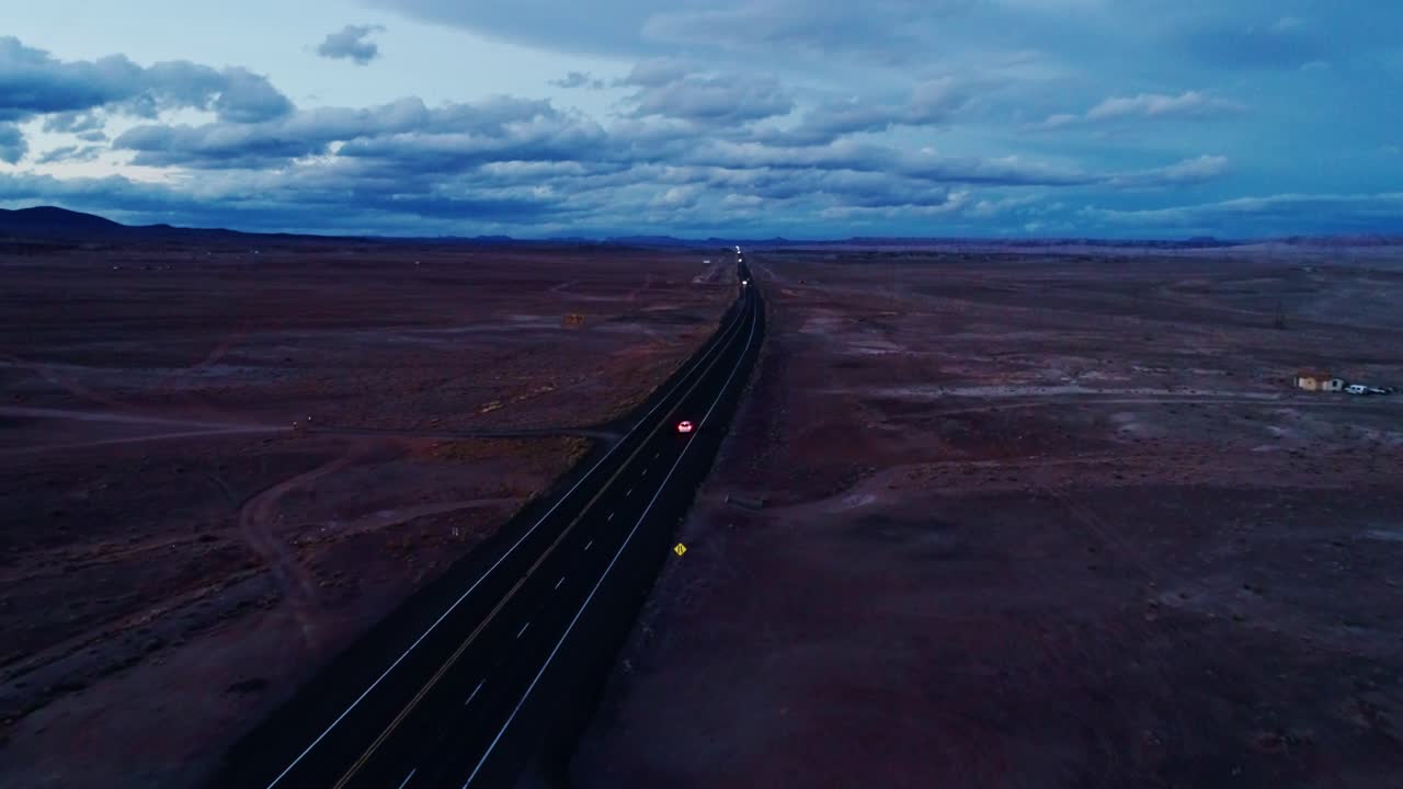 Remote highway vanishing into desert dusk with red car lights in distance, drone