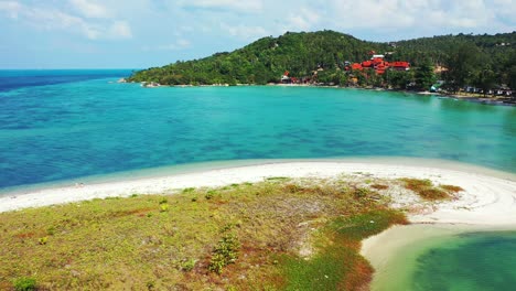 Calm-turquoise-sea,-small-sandbank-with-tropical-vegetation-and-coast-with-hills-and-palms-in-the-background,-Aerial-view