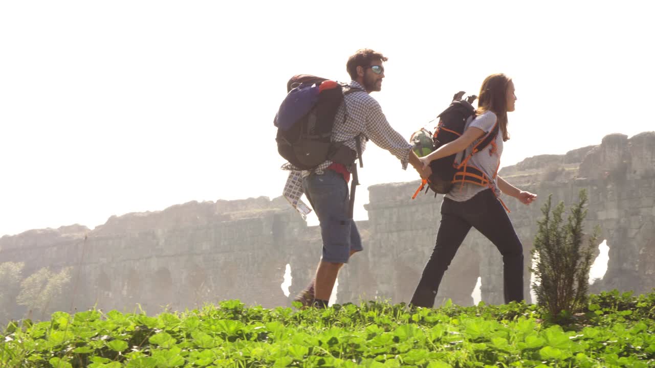 Giovane coppia di turisti zaino in spalla che cammina mano nella mano verso gli archi dell'acquedotto romano nelle rovine del Parco degli Acquedotti a Roma in una romantica alba nebbiosa, con chitarra e sacco a pelo, in slow motion.