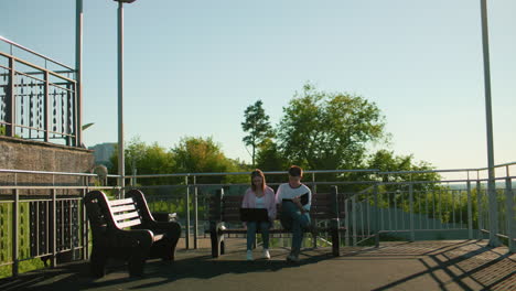 high school students study outdoors; girl with laptop on legs, boy taking notes, amidst greenery and light poles enhancing the peaceful academic setting