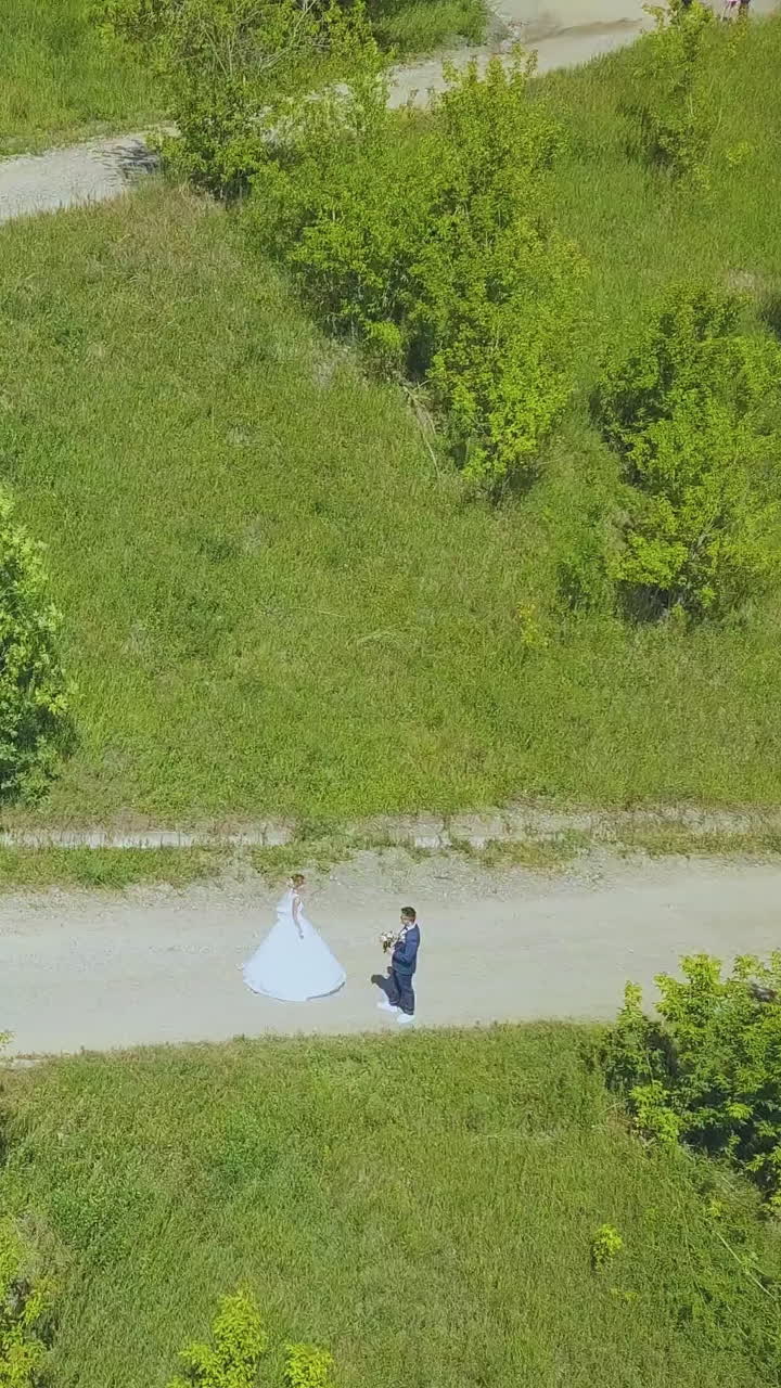 happy bride spins near groom on hairpin curves of path on lush green hill on nice sunny day upper view