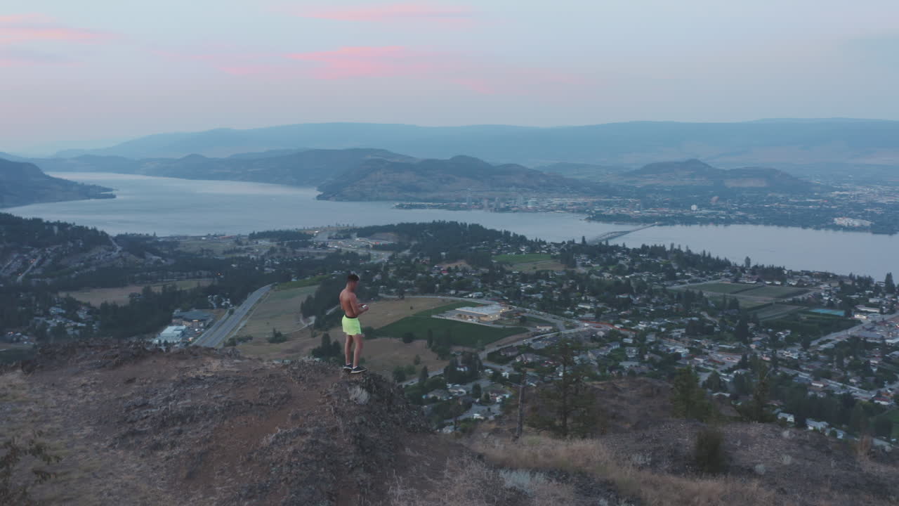 Man stands at top of peak to take photo of mountains, city and lake below