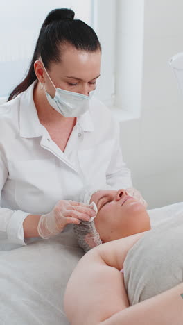 cosmetologist doctor wipes woman face and neck cleaning procedure with cotton wipes in beauty clinic