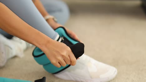 a person fastens ankle weights over sneakers on a carpeted floor, preparing for a workout session