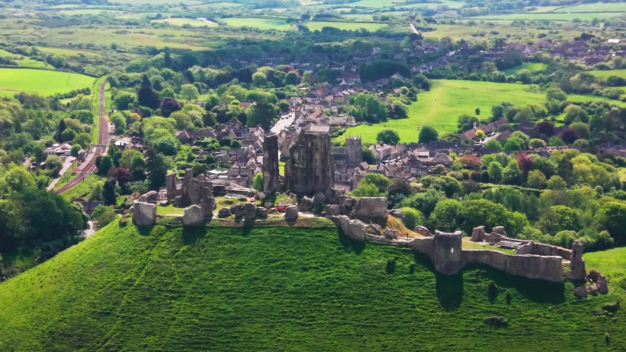 Droneoptagelse af Corfe Castle-ruinerne, der står på en grøn bakke over landsbyen Corfe Castle i Dorset, England. Billedet viser de historiske middelalderlige slotsruiner og landsbygader.
