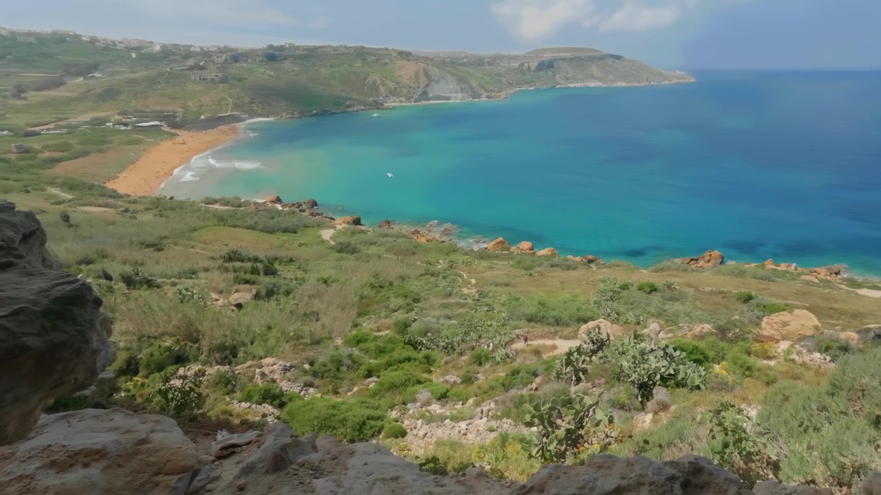 Un primo piano roccioso incornicia la Grotta di Mixta a Gozo, che a sua volta fa da cornice al vivace mare turchese e alla sabbiosa spiaggia di Ramla sottostante. Ideale per promozioni di viaggio o documentari naturalistici.