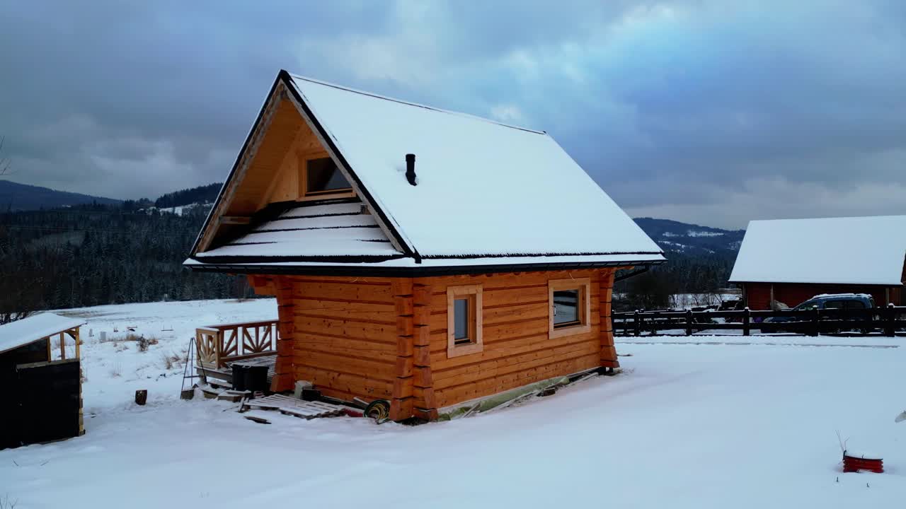 En håndlaget fjellhytte innrammet av snødekt terreng og skogkledde topper. Drone cinematic dolly-out og zoom-in bevegelse forsterker den dramatiske vinterstemningen