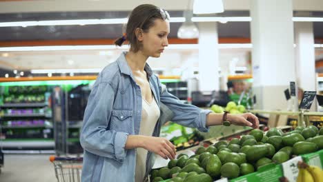 Vegetable-and-fruits-department-in-the-grocery-store.-Pretty-girl-in-casual-selecting-green-avocado.-Handheld-footage.-Side-view