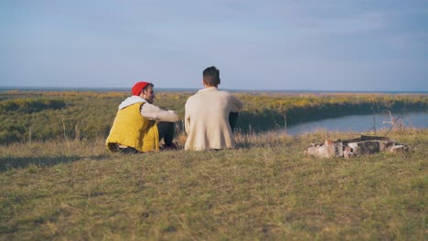 young men spend time sitting on hilly river bank at campsite