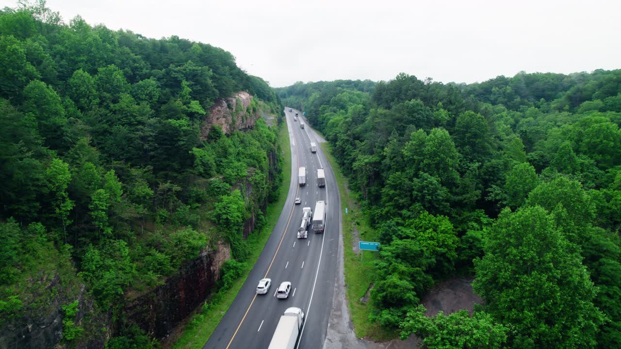 Aerial footage of tanker and semi trucks driving along a lush Tennessee highway carved through green forest cliffs in summer.