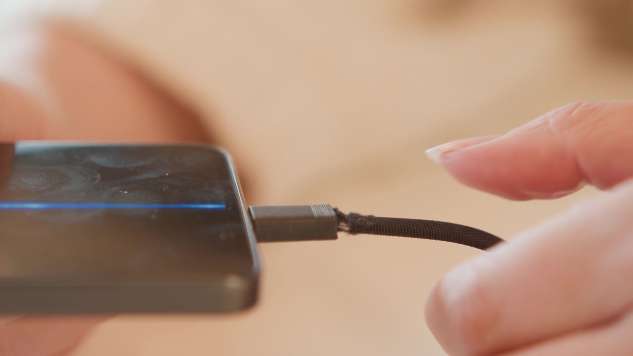 Side view of hands pushing braided charging cable into smartphone port, with visible fingertip pressure, connector alignment, and tactile contact under warm indoor light, with smartphone, cable