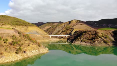 calm emerald water of lake reflecting bridge on rocky shore of rural landscape near mountain village, aerial view