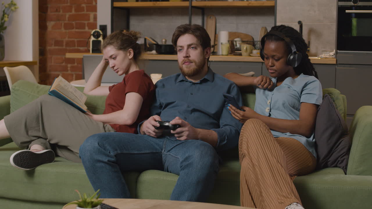 три товарища читают книгу, играют в видеоигры и слушают музыку со смартфона, сидя дома на диване