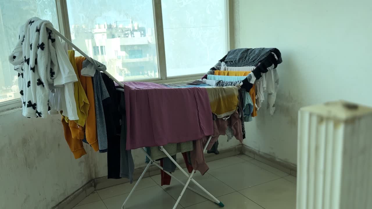 Interior shot of a foldable drying rack filled with colorful laundry placed beside large windows overlooking residential buildings. Daylight scene capturing everyday home drying routine