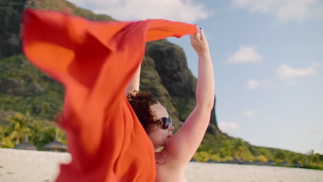 a woman enjoys a sunny beach day, lifting a red scarf into the air