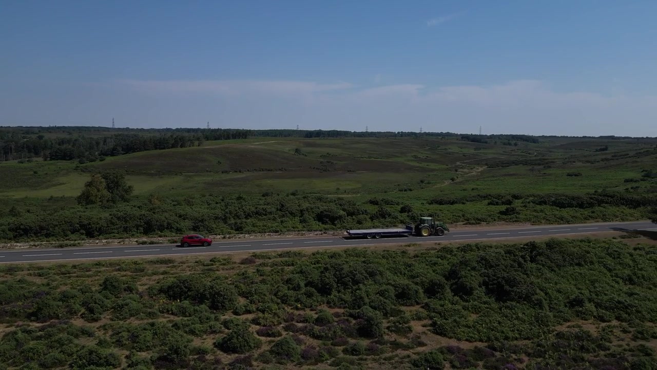 Aerial drone shot of tractor towing trailer along rural road through heathland in New Forest, Hampshire, England with surrounding woodland beneath clear daytime sky