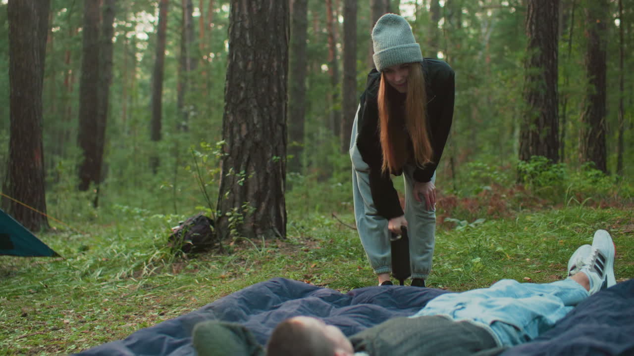 Young woman pumps air into tent bag while man lays on it, surrounded by forest trees and pitched tent in background, showcasing outdoor teamwork