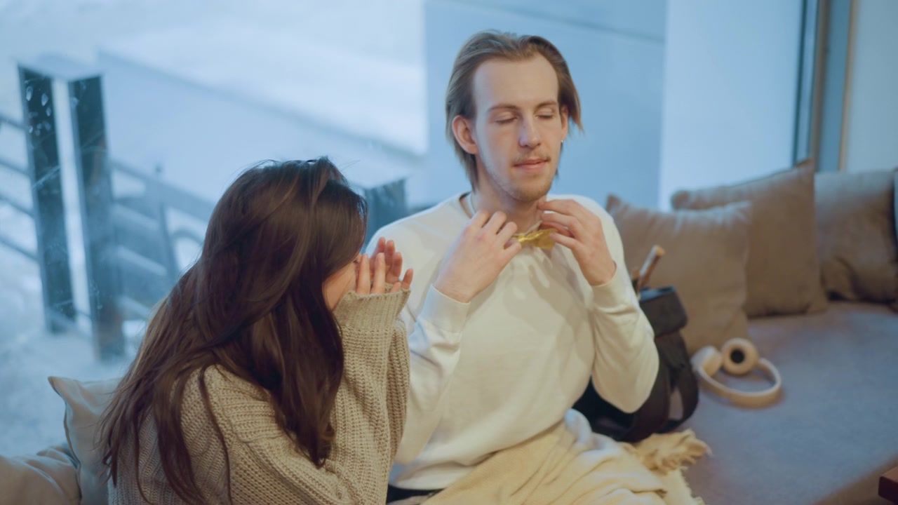 Close up of young woman and man sitting together indoors girl watching as boy adjusts his yellow bow tie. scene features soft lighting with visible objects like headphones on couch cushion