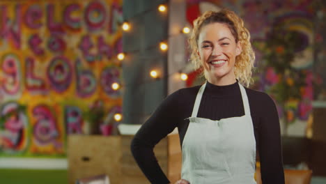 Portrait-Of-Smiling-Female-Server-Or-Owner-Wearing-Apron-Walks-Into-Focus-In-Cool-Bar-Or-Club