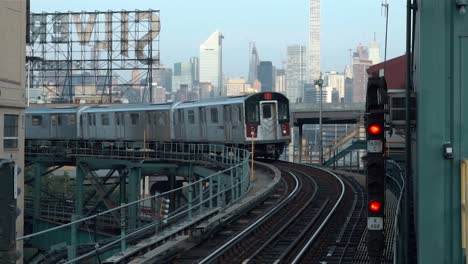 Einfahrender-Zug-An-Der-U-bahnstation-Queensboro-Plaza-Mit-Der-Skyline-Von-Manhattan-Und-Den-Sivercup-Studios-Im-Hintergrund,-Gefilmt-An-Einem-Sonnigen-Morgen-In-New-York-City