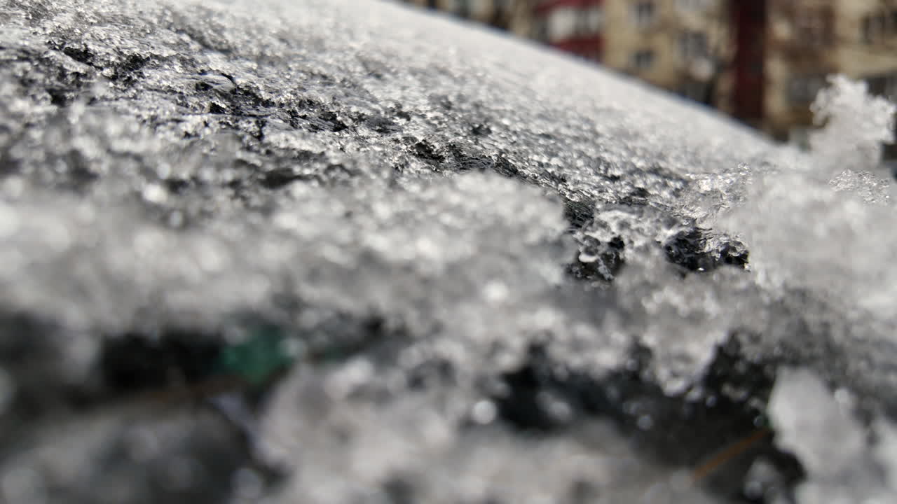 Primer plano de cristales de hielo congelados cubriendo el parabrisas de un coche, con edificios urbanos borrosos al fondo durante condiciones de frío invernal.