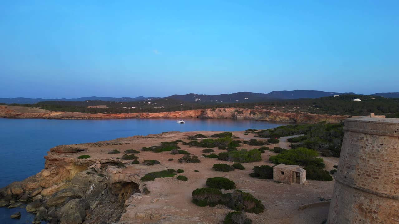 Carefree woman in pink dress Yoga tree pose enjoying freedom and breathtaking view on Ibiza island near Torre des Savinar at sunset. Lovely aerial view flight ascending drone